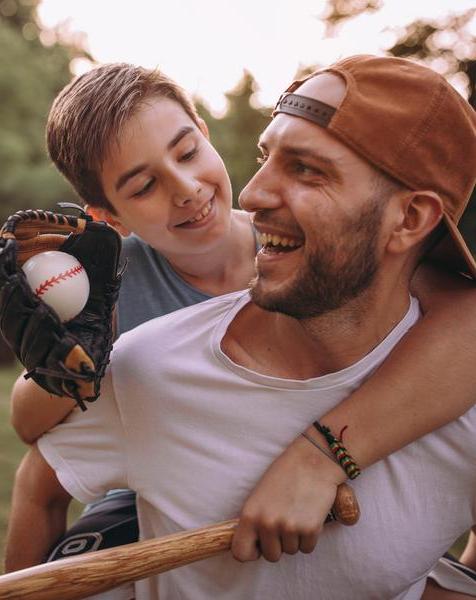 小孩坐在男人的背上，拿着棒球棒、手套和球.
